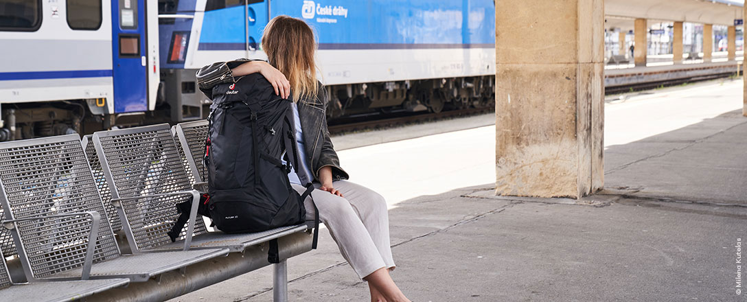 Person sitzt mit Rucksack am Bahnsteig, im Hintergrund ein blauer Zug