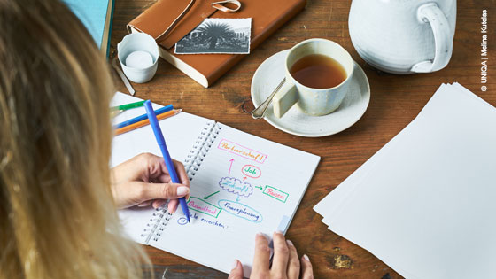 Frau sitzt am Tisch und notiert Gedanken zu ihren Träumen und Zielen in ein Notizbuch