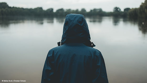 Person steht einsam mit Regenjacke vor einem See 