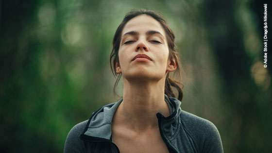 Frau ist im Wald, hebt den Kopf leicht nach oben und hat die Augen geschlossen
