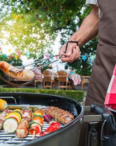 Mann steht bei offenem Kugelgrill und hält Grillzange mit Wurst