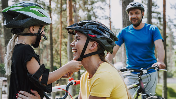 Kind schließt Helm von lachender Frau, im Hintergrund ein Mann